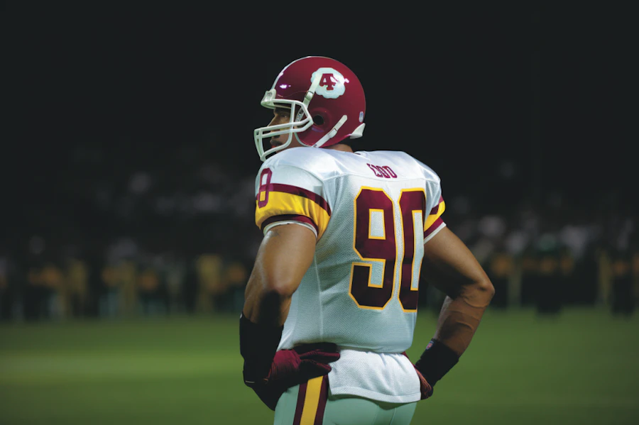 A football player in a white jersey stands with hands on hips, looking off toward the sideline at night